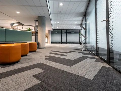 Finished office floor with patterned carpet tile, orange upholstered stools, a concrete column, recessed ceiling lights and glass fronted rooms with frosted film