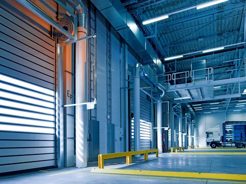 Warehouse loading area with a row of high speed roll-up doors, yellow bollard rails on a concrete floor, a steel walkway overhead and a truck parked at the far end