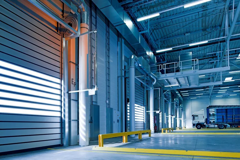 Warehouse loading area with a row of high speed roll-up doors, yellow bollard rails on a concrete floor, a steel walkway overhead and a truck parked at the far end
