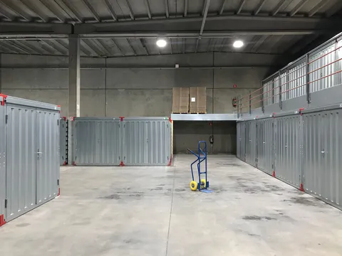 Warehouse bay with grey steel storage units on a concrete floor, a mezzanine walkway with orange handrails along the right wall and a blue hand truck in the middle