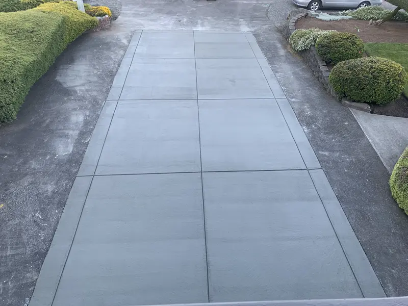 New concrete driveway with a broom finish and straight control joints, between shrubs and a retaining wall