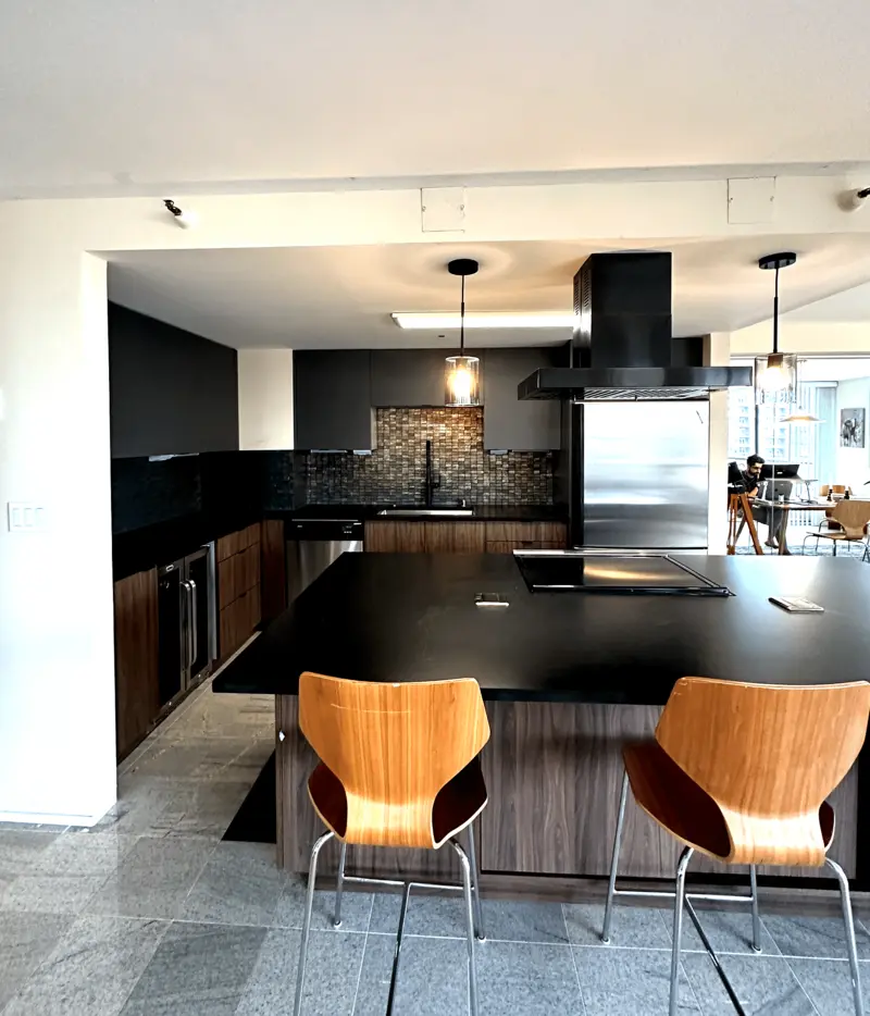 Open condo kitchen with a black island, wood-grain cabinets, a black range hood, pendant lights and bar stools on a stone tile floor