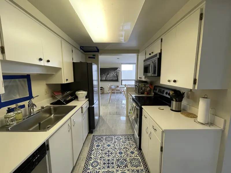 Narrow white galley kitchen with a fluorescent ceiling panel, a patterned runner and a gray stone tile floor