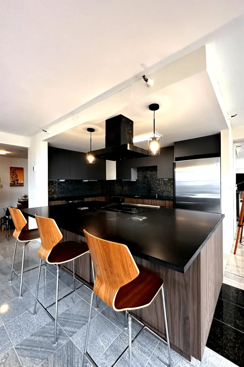 Black island with a cooktop and three wood bar stools under a black range hood and pendant lights