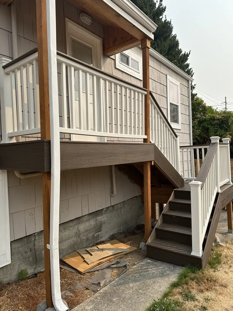 Covered upper landing on wood posts with a white railing and a stair down the side of the house
