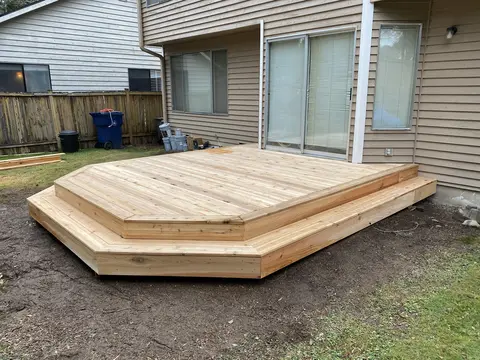 New low platform deck of light wood with angled corners and a step around the front edge, outside a sliding door