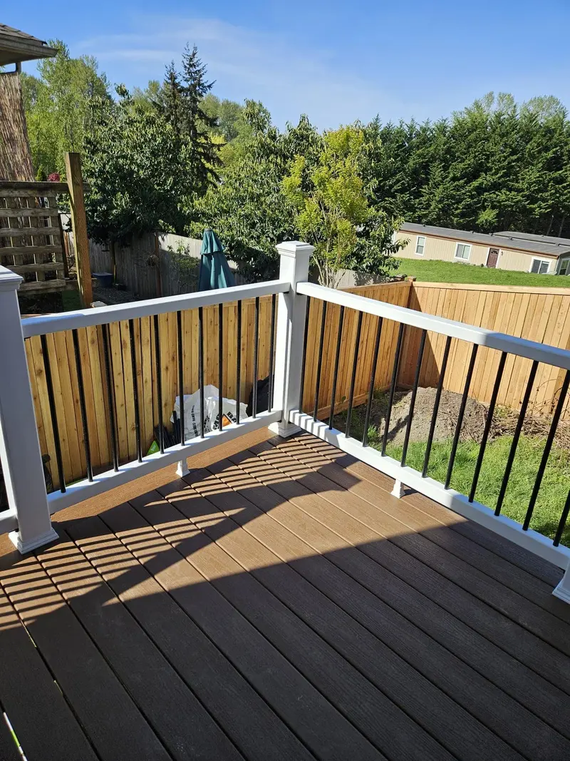 Deck corner with brown boards and a white railing, a wood fence and trees beyond