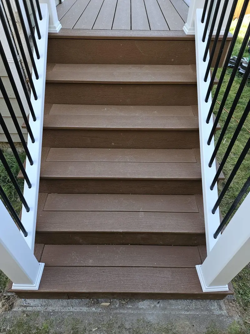 Looking down a deck stair with brown treads between white railing posts and black balusters