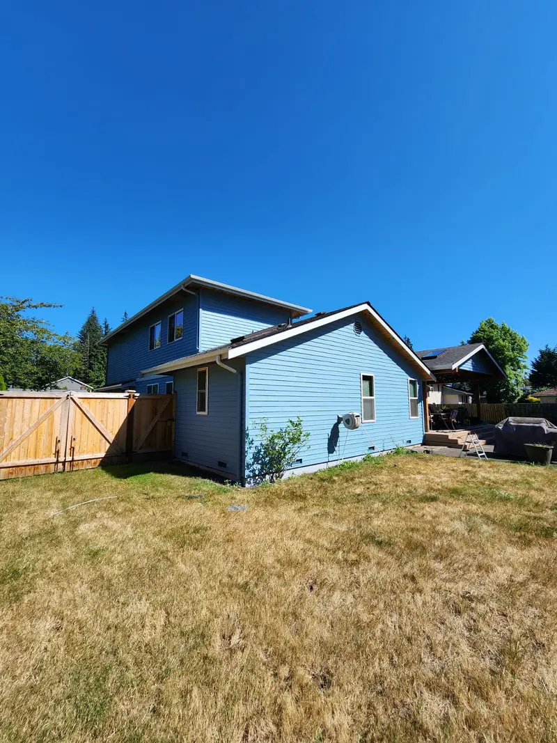 Side of the blue house beside a wood fence and gate, seen across a dry lawn