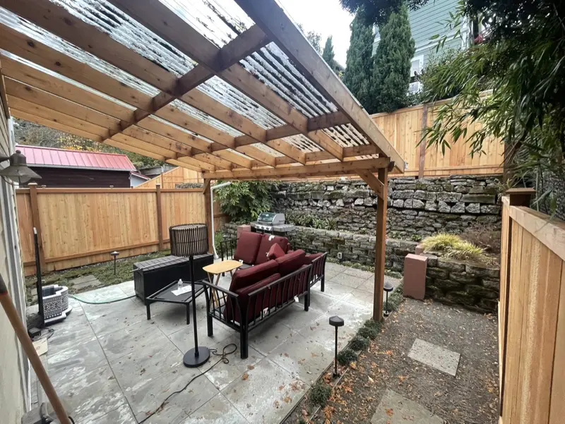Paved patio with red outdoor sofas under a wood pergola, enclosed by new cedar fencing and stone retaining walls