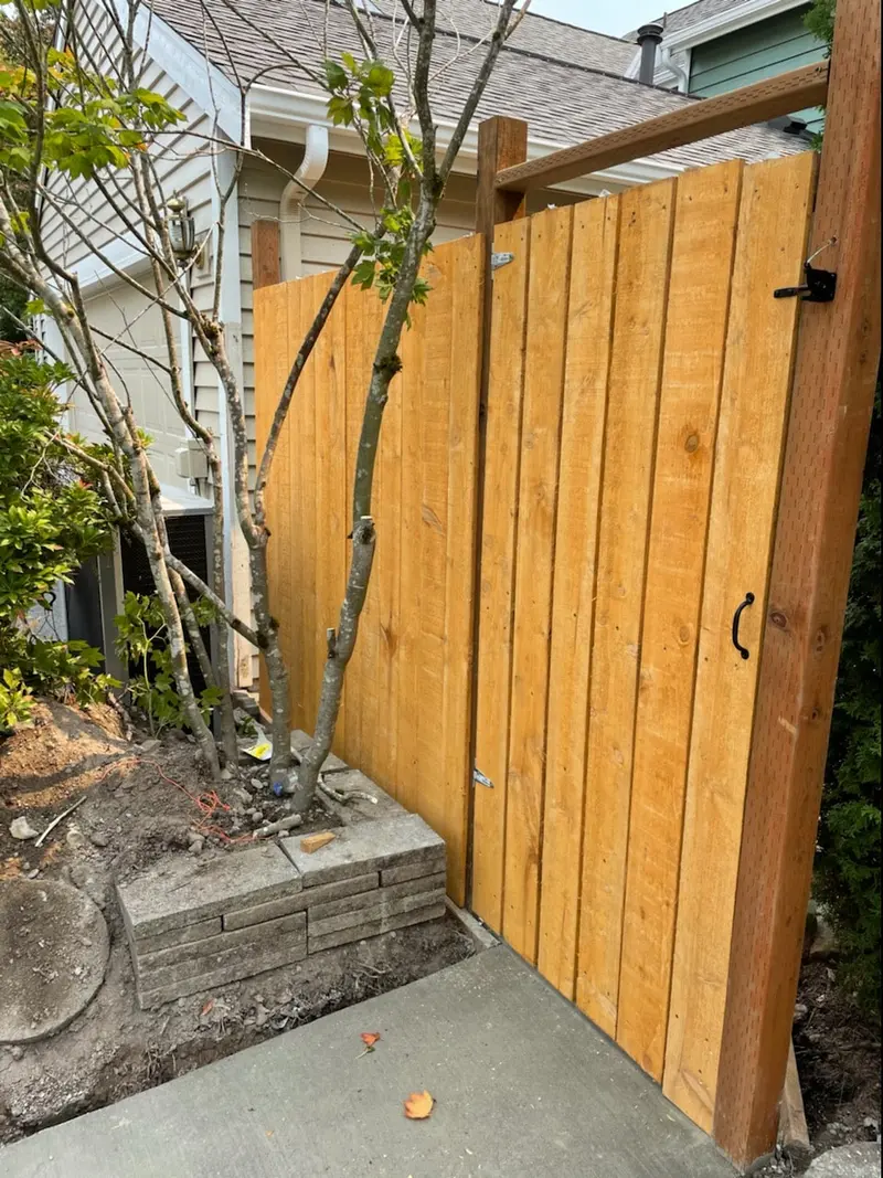 Tall wood gate with a black pull handle and latch, hung on posts beside a small tree and a block planter