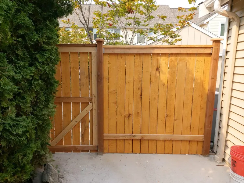 Wide wood fence section with a framed gate on the left, standing across a concrete pad beside a house