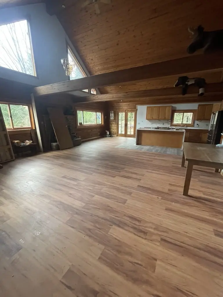 Great room with a new warm wood-look plank floor under exposed beams, looking toward the kitchen