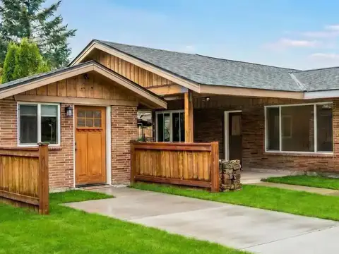 Brick house whose former garage wing now has a wood front door and a window, under a cedar board and batten gable, with the concrete driveway leading up to the door