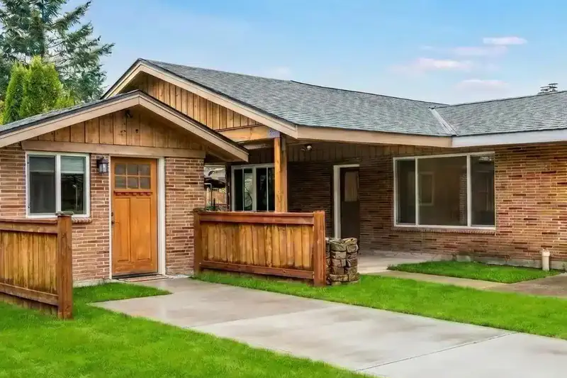 Brick house whose former garage wing now has a wood front door and a window, under a cedar board and batten gable, with the concrete driveway leading up to the door