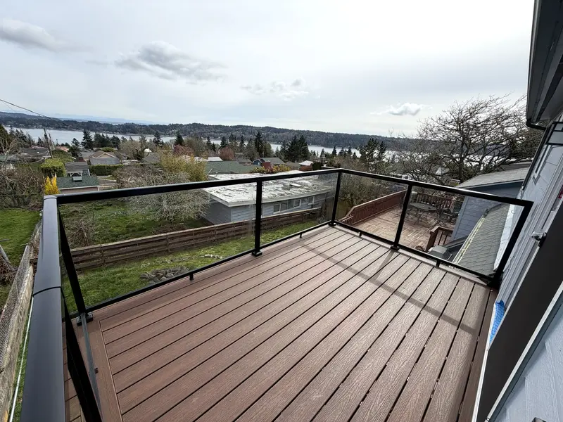 Upper deck with brown composite boards and black-framed glass railing looking over rooftops to water and distant hills