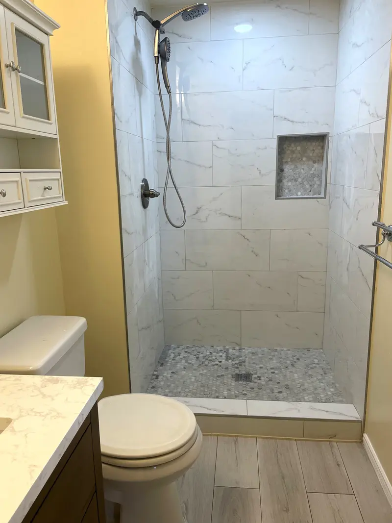 Walk-in shower with large marble-look wall tile, a pebble mosaic floor and a handheld shower on a slide bar