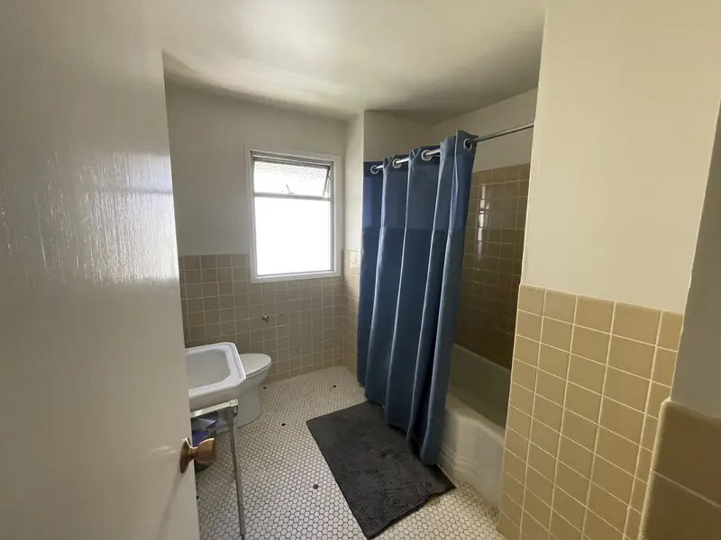 Guest bathroom before the remodel with tan wall tile, a blue shower curtain and a pedestal sink