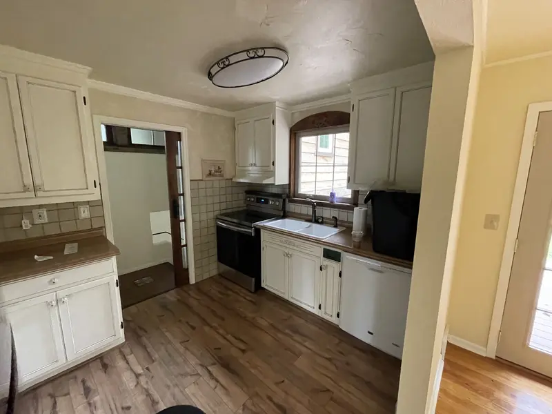 Older kitchen with white painted cabinets, a tile backsplash, a wood floor and a gas range under a ceiling light