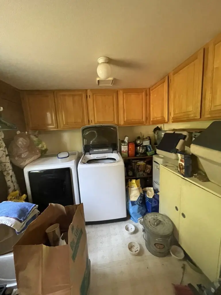 Laundry room before the remodel, crowded with boxes beside the washer and dryer under wood cabinets