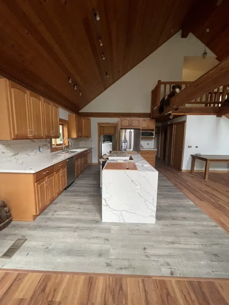 Log home kitchen with a white waterfall island on a gray plank floor under a vaulted cedar ceiling