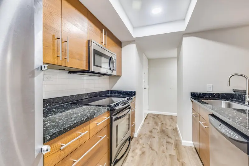 Galley kitchen with wood cabinets, dark granite counters, stainless appliances and a light plank floor