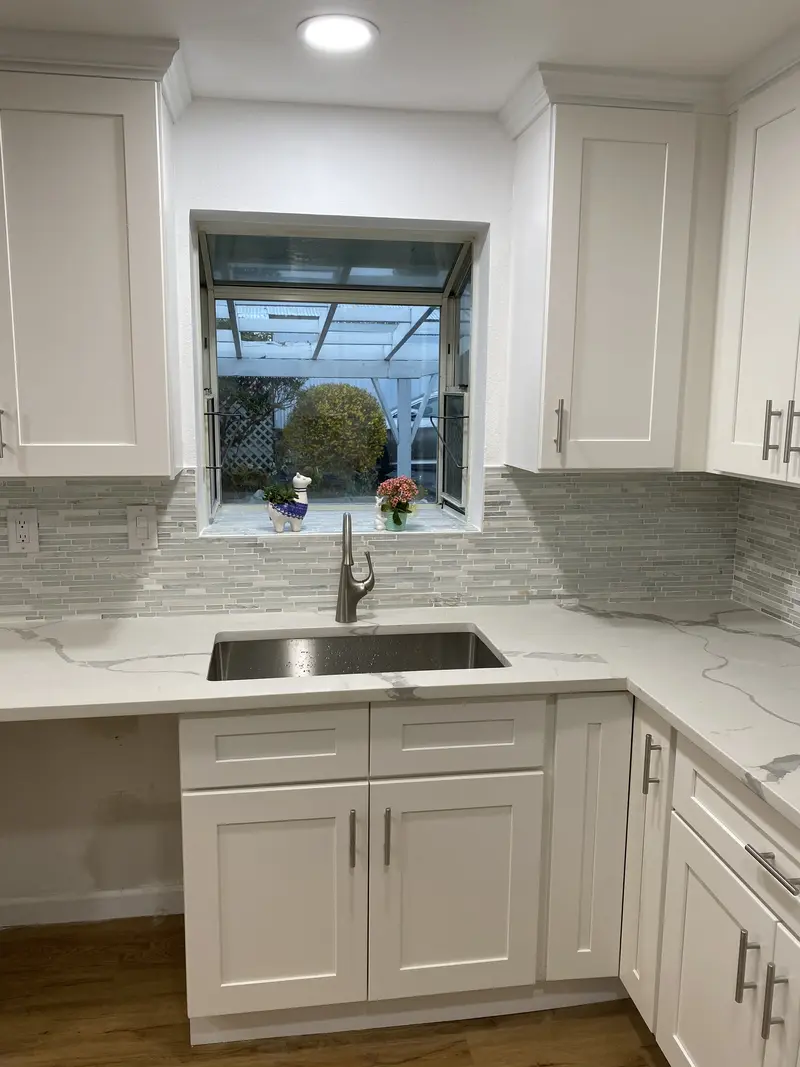White shaker kitchen with a stainless sink under a window and a gray glass mosaic backsplash