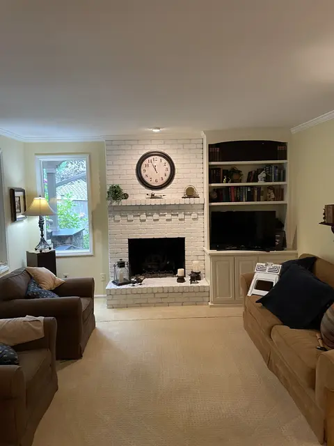 Living room with the brick fireplace and hearth painted white, a wall clock above and built-in shelves to the right