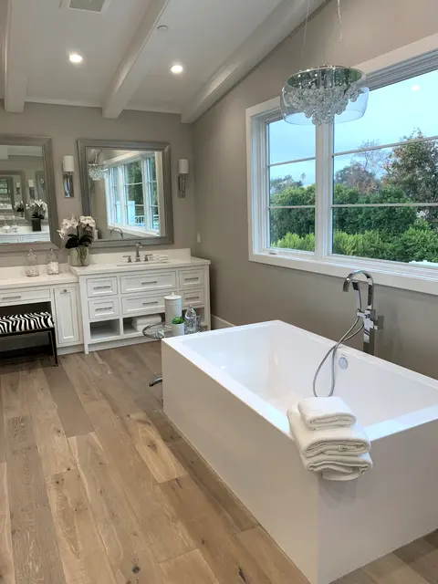 Freestanding rectangular tub with a floor-mounted filler under a wide window, below a crystal chandelier and exposed ceiling beams