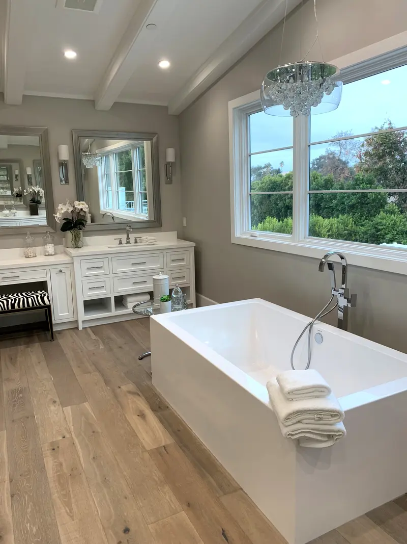 Freestanding rectangular tub with a floor-mounted filler under a wide window, below a crystal chandelier and exposed ceiling beams