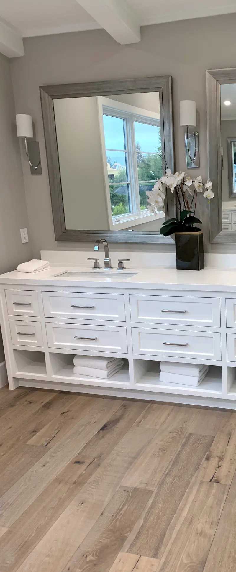 White vanity with drawers, an undermount sink and open towel shelves below a framed mirror and wall sconces