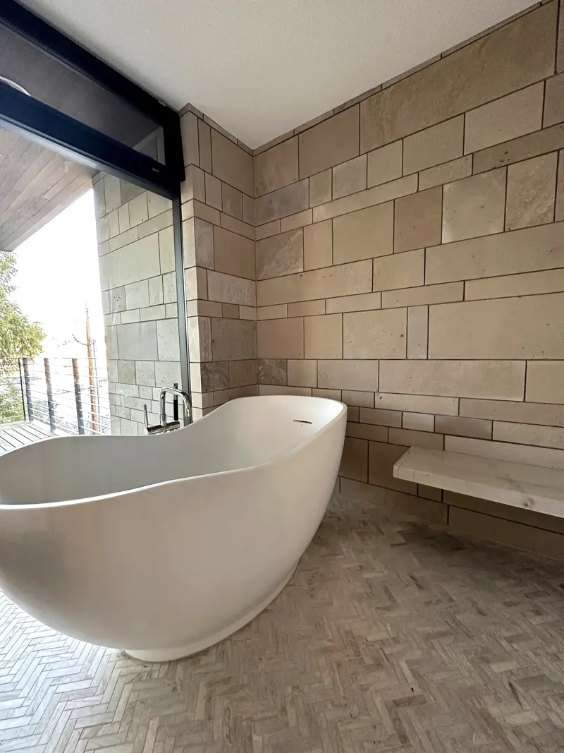 White freestanding tub on a herringbone floor beside a tall window, with stone-look tile walls and a floating bench