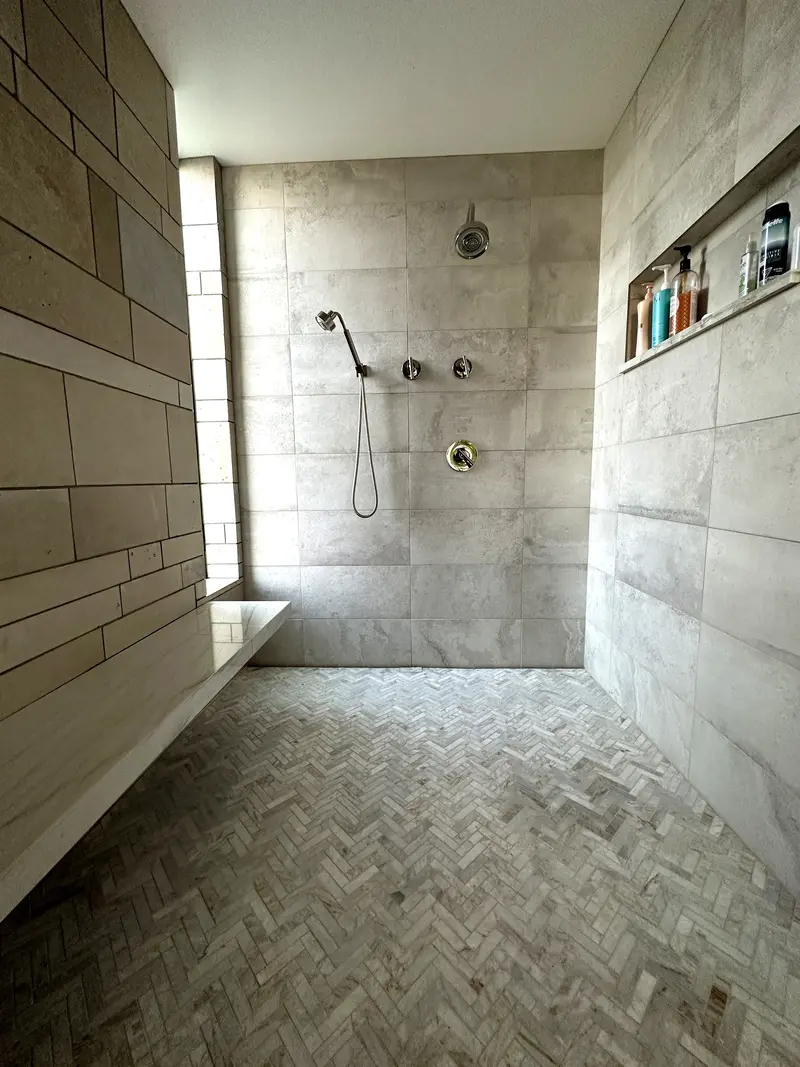 Open curbless shower with stone-look wall tile, a long recessed niche, a wall shower head and a handheld spray over a herringbone floor