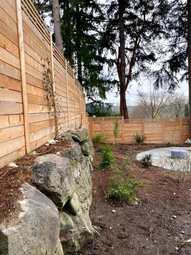 Horizontal cedar fence running along a boulder retaining wall, with new planting beds and a round patio beyond