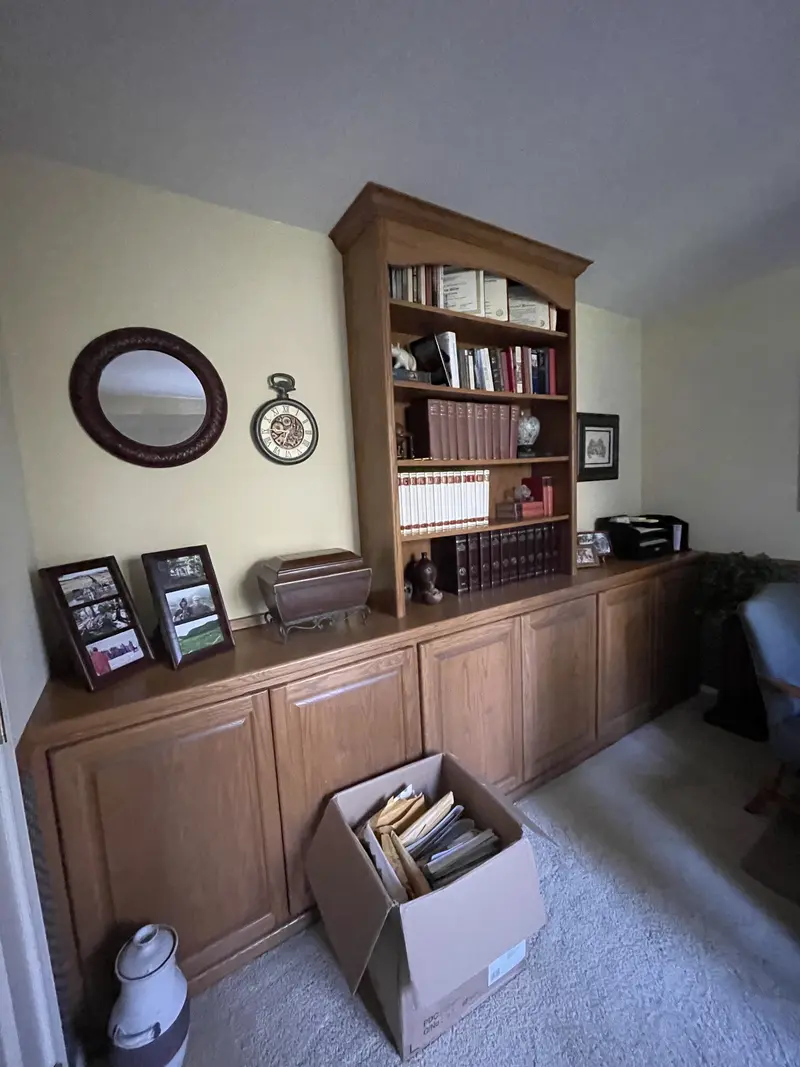 Office wall with stained wood base cabinets and a tall stained bookcase with books, a box of files on the floor