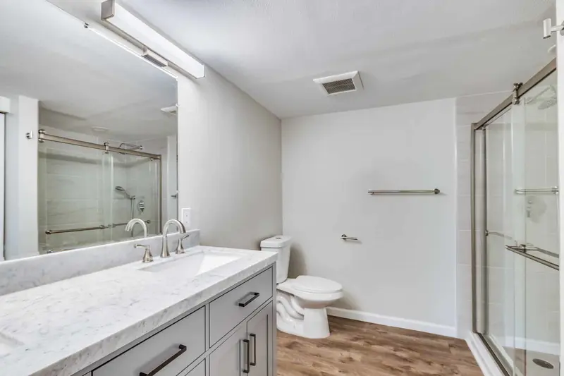 Bathroom with a gray shaker vanity, white counter, wide mirror and a glass shower door