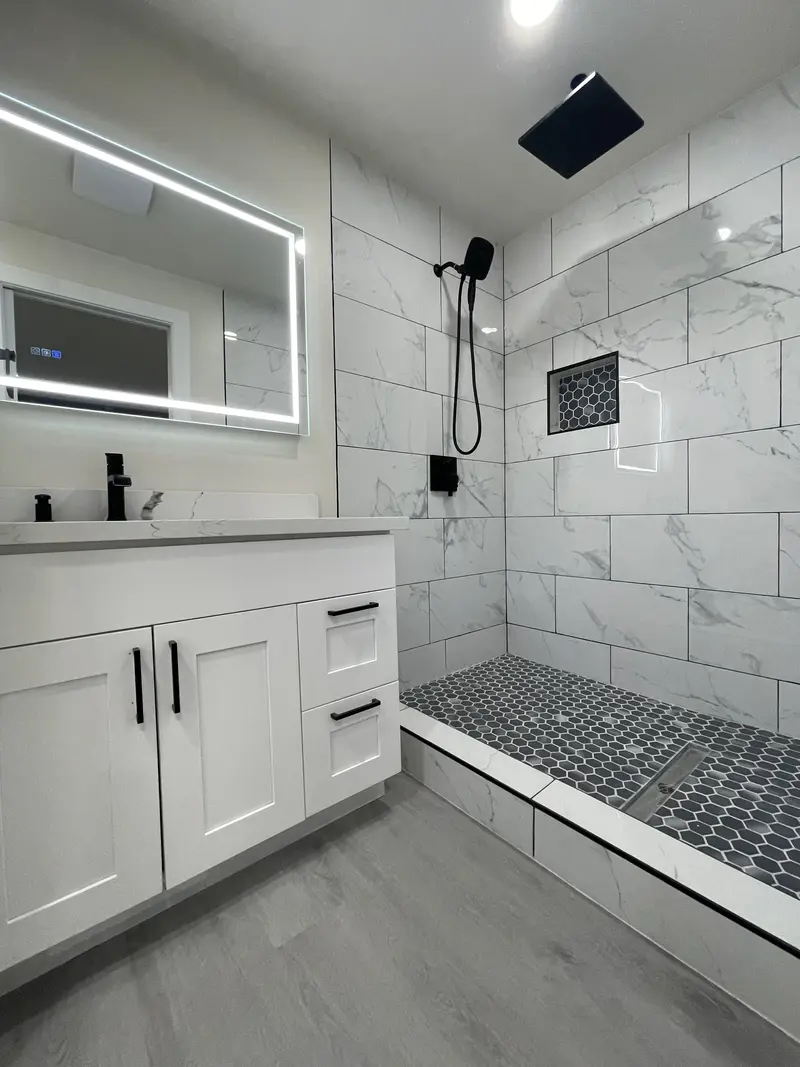 Walk-in shower with large-format marble-look tile, black rain head, pebble floor and a lit mirror over the vanity
