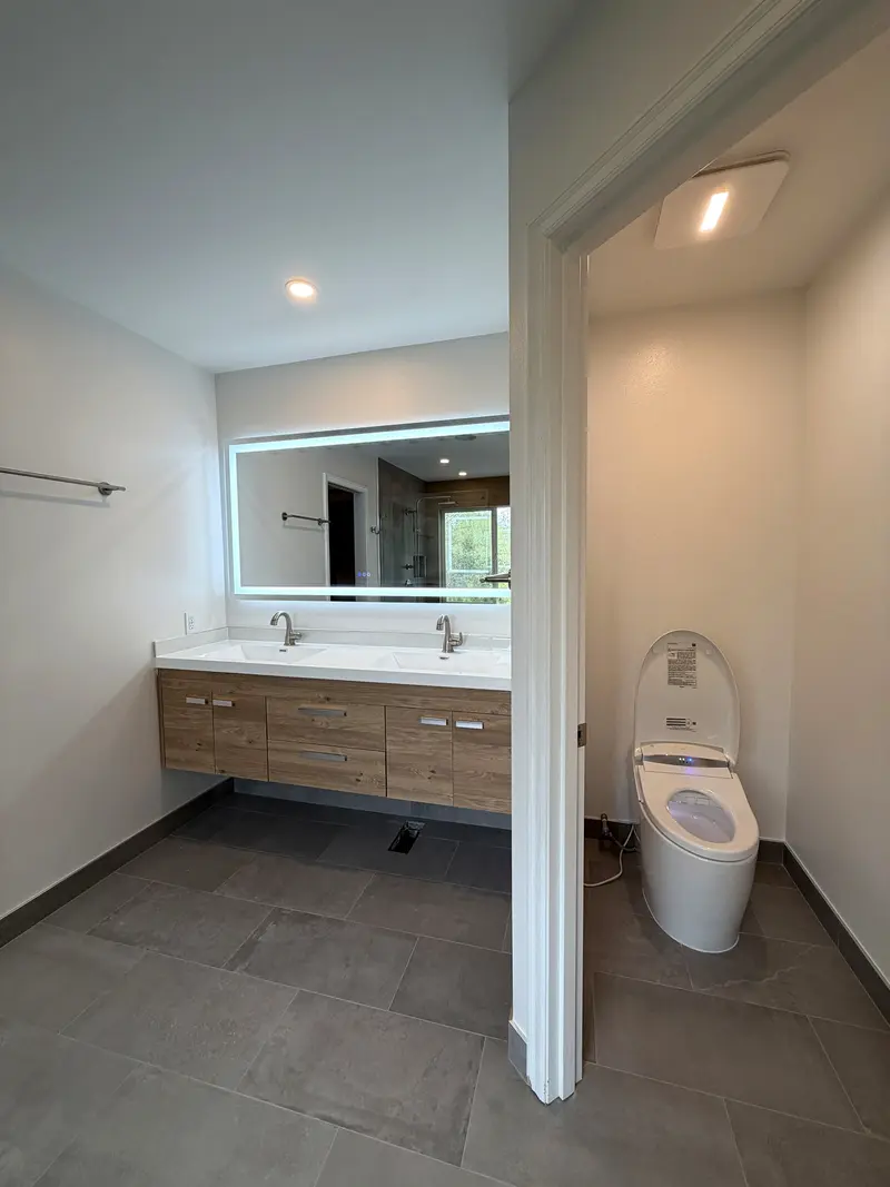 Floating wood double vanity under a lighted mirror, beside a doorway into a separate toilet room