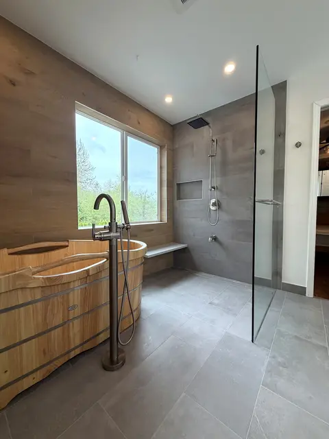 Wooden soaking tub with a floor-mounted filler by a window, beside a walk-in shower behind a single glass panel