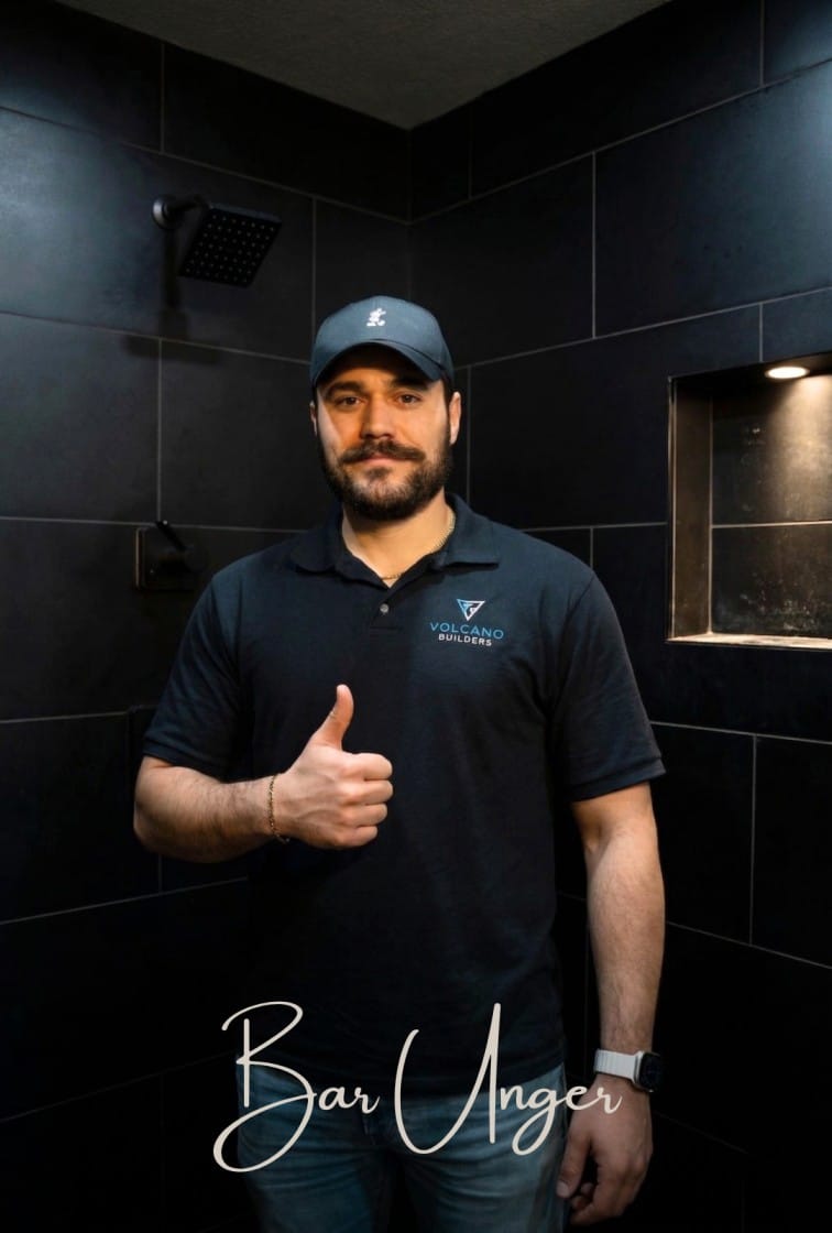 Bar Unger owner of Volcano Builders standing inside a completed dark tile bathroom remodel with rain shower head and built in niche in Washington State